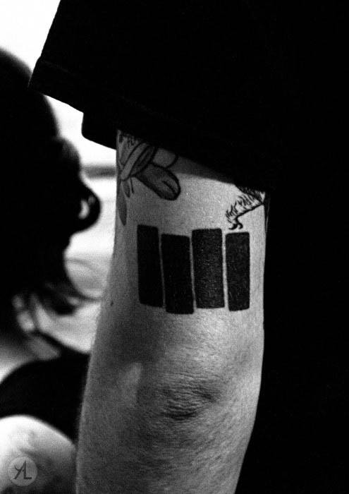 Classic Black Flag bars tattooed on the tricep, with a small burning cigarette illustrated above one of the bars for added rebellious flair.