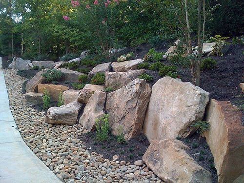 Sloped landscape featuring large natural boulders, small plants, and a pebble-lined border for a rugged and low-maintenance design