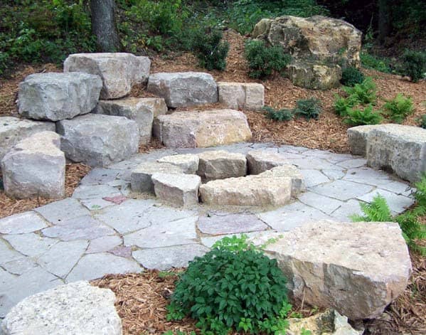 Stone fire pit surrounded by large rocks and greenery on a stone patio
