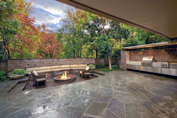 Outdoor patio with stone flooring, a circular fire pit, seating, and a built-in grill, surrounded by trees