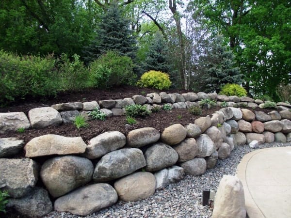 Tiered rock retaining wall with mulch, plants, and evergreen trees along a gravel pathway.