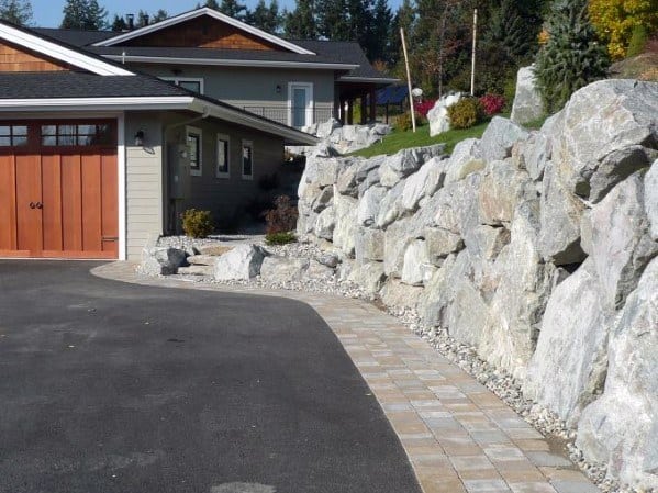 Asphalt driveway with paver edging bordered by a large stone retaining wall near a modern house.