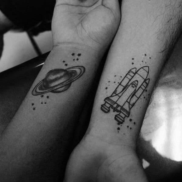 Matching wrist tattoos of a planet and a rocket surrounded by stars, symbolizing a love that’s out of this world and destined to explore together.