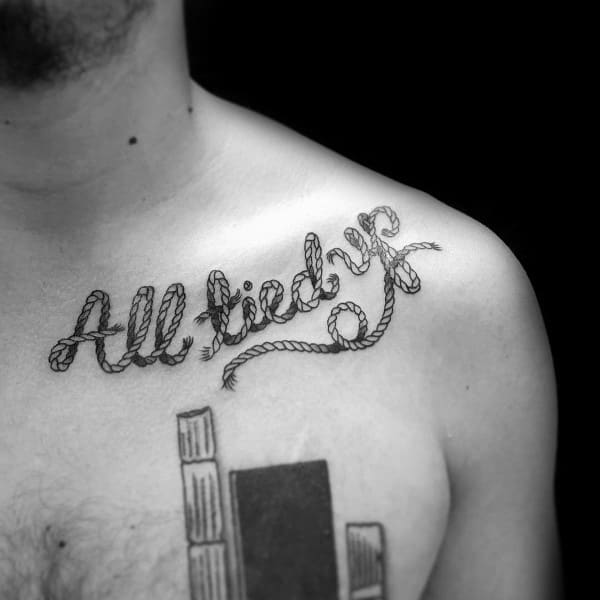 Black and grey chest tattoo featuring the phrase "All tied up" in rope-style lettering, designed with fine details and a Western-inspired theme.