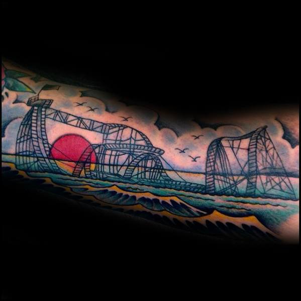Colorful roller coaster tattoo with a seaside theme, featuring waves, a red sunset, birds, and a dramatic sky in the background.
