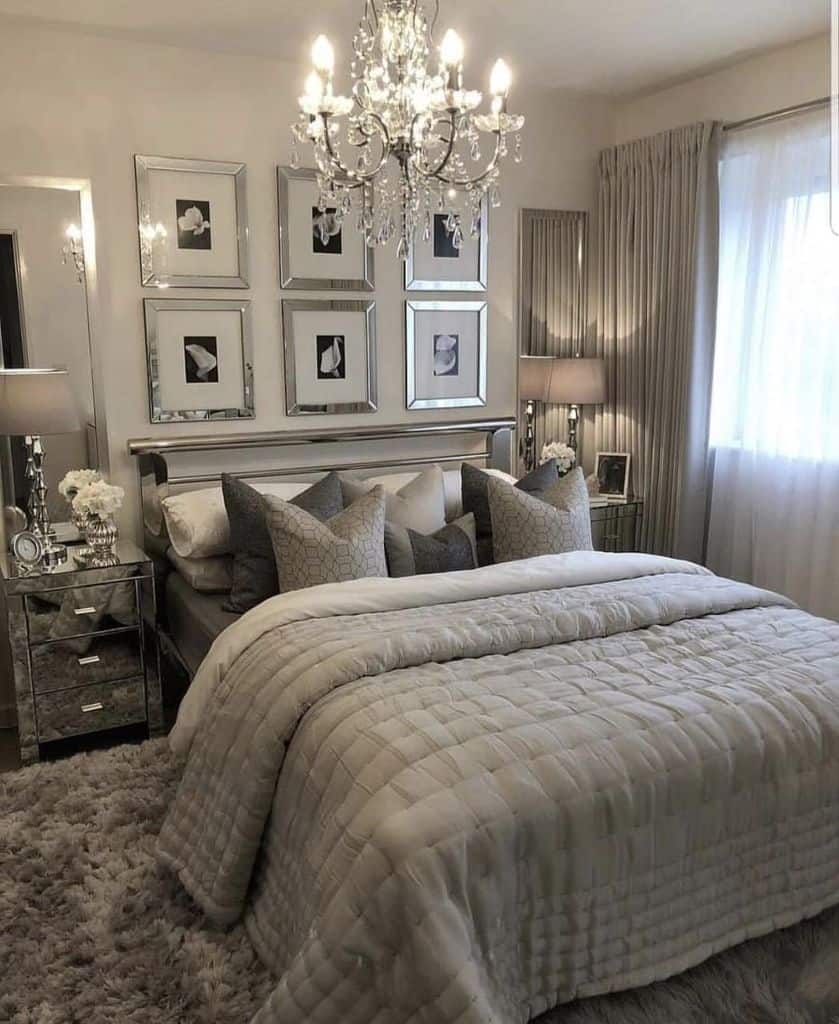 Elegant bedroom with a chandelier, plush bedding, mirrored furniture, and framed art on the wall, featuring neutral tones throughout