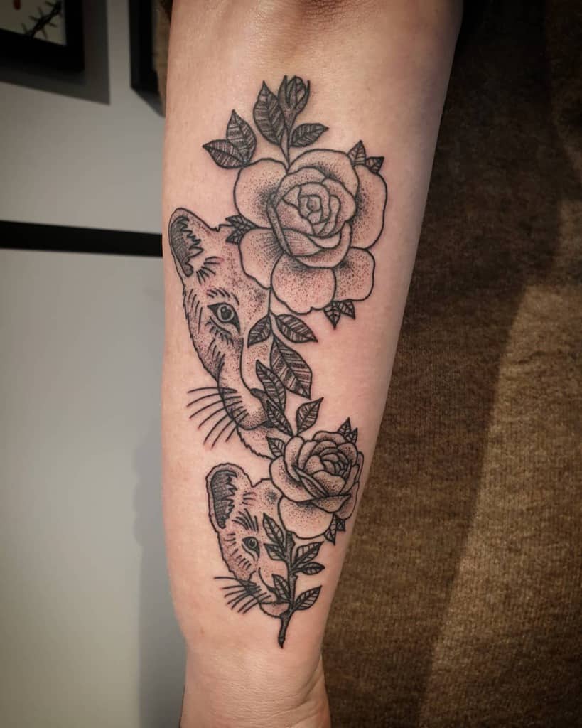 Black and grey forearm tattoo featuring two lionesses intertwined with detailed roses and leaves, created with bold linework and dot shading.
