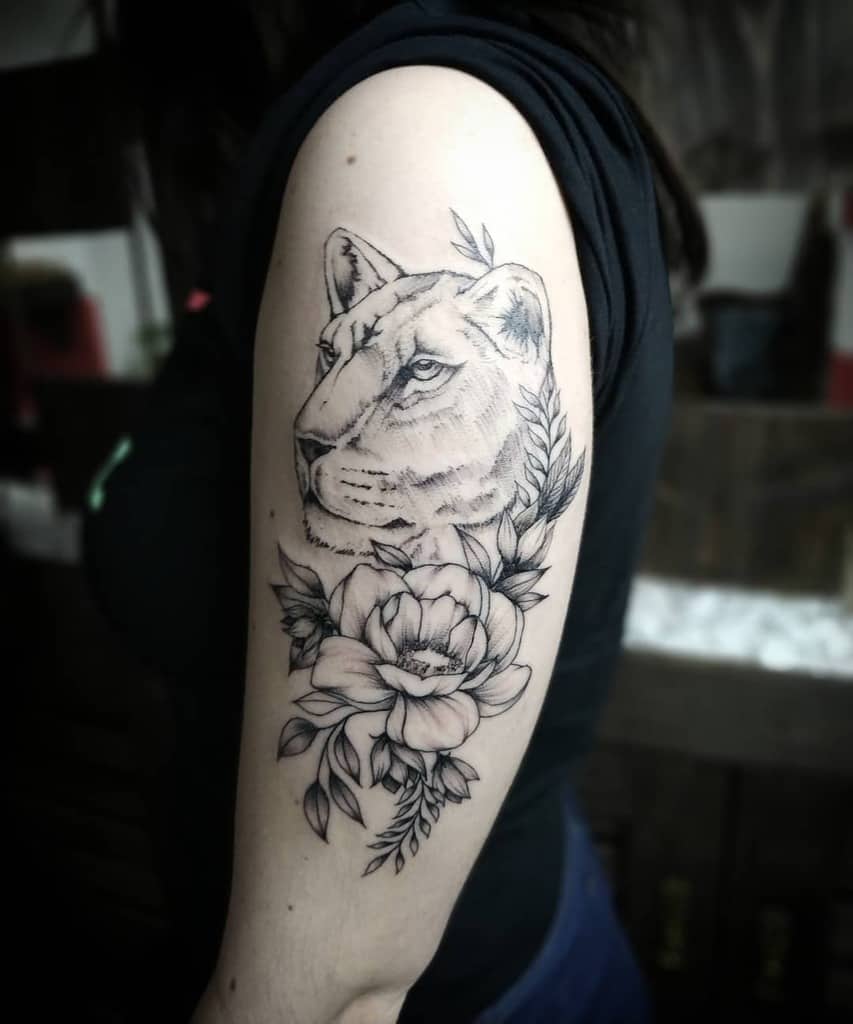 Elegant black and grey lioness tattoo on the upper arm, featuring fine shading, floral elements, and leaves for a graceful yet powerful look.