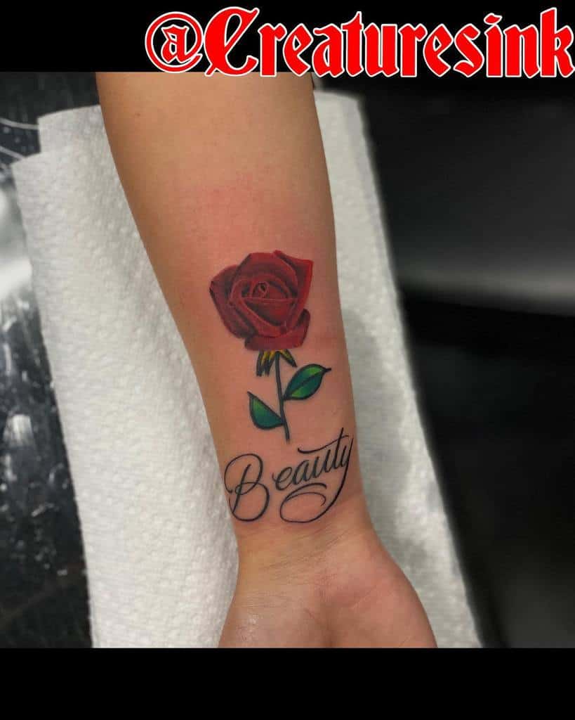 A tattoo of a red rose with green leaves graces the wrist above the word "Beauty" on an arm