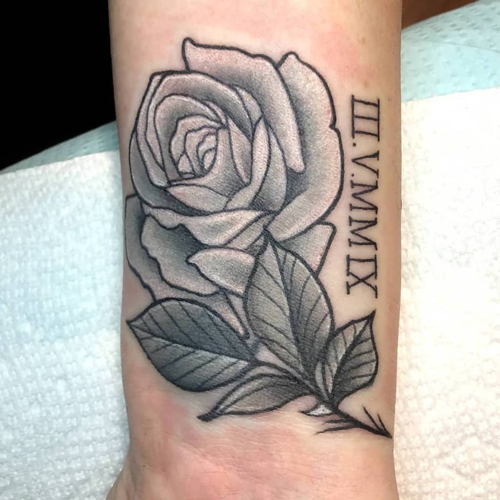 Tattoo of a rose and leaves with Roman numerals "III.V.MMIX" gracefully wraps around the wrist