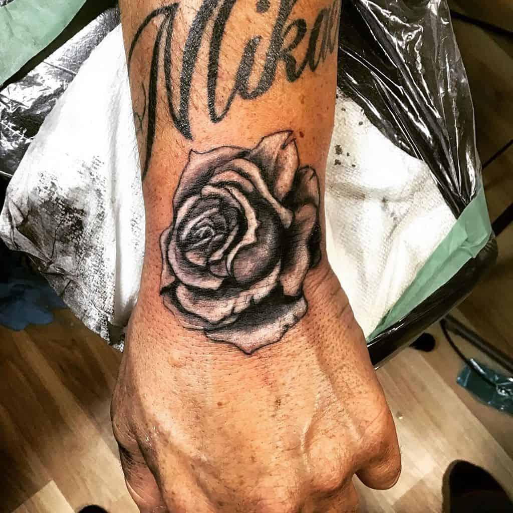 A detailed black and gray rose tattoo elegantly blooms across a person's hand, seamlessly blending into script lettering that graces the wrist