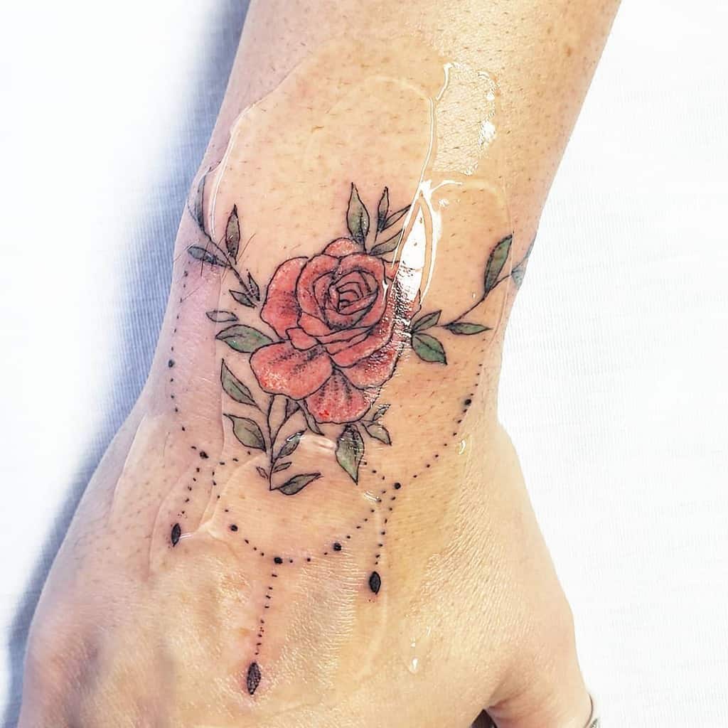 Delicate rose tattoo with soft red and green tones, placed on the back of the hand, accented with decorative dotted lines
