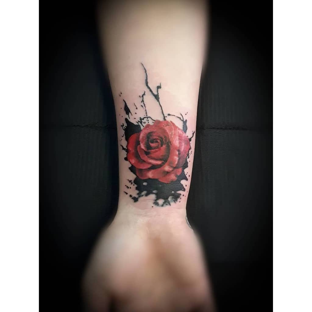 A red rose tattoo with black ink splatters on a wrist
