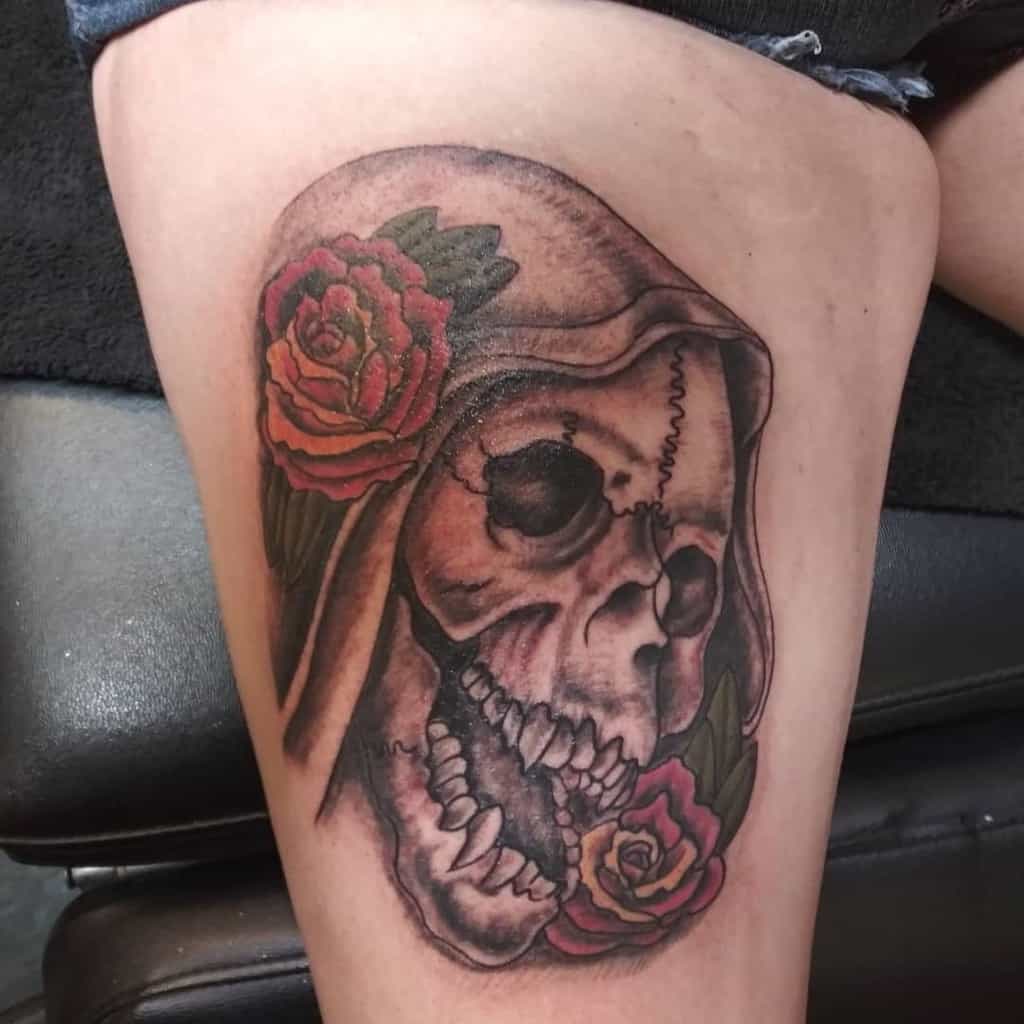 Thigh tattoo of a hooded skull with red roses, blending dark realism with floral elements for a striking and eerie design.