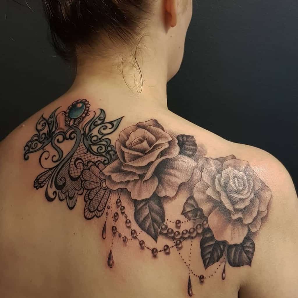 Elegant shoulder tattoo featuring realistic roses, intricate lace details, beaded chains, and a decorative gemstone ornament.