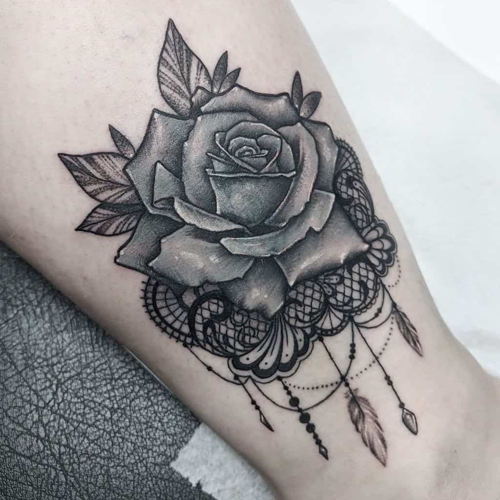 Black and grey tattoo featuring a realistic rose with intricate lace patterns, delicate beaded chains, and feather charms on the leg.