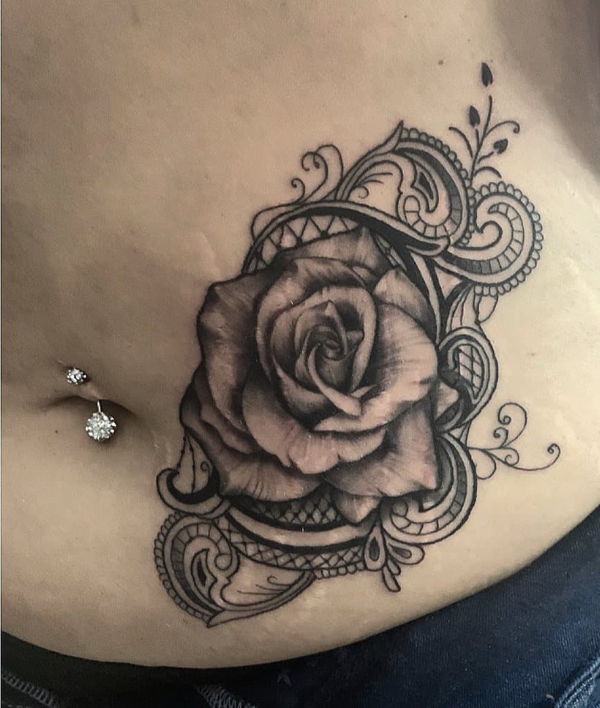 Black and grey tattoo on the lower abdomen featuring a realistic rose with intricate lace patterns and ornamental filigree details.