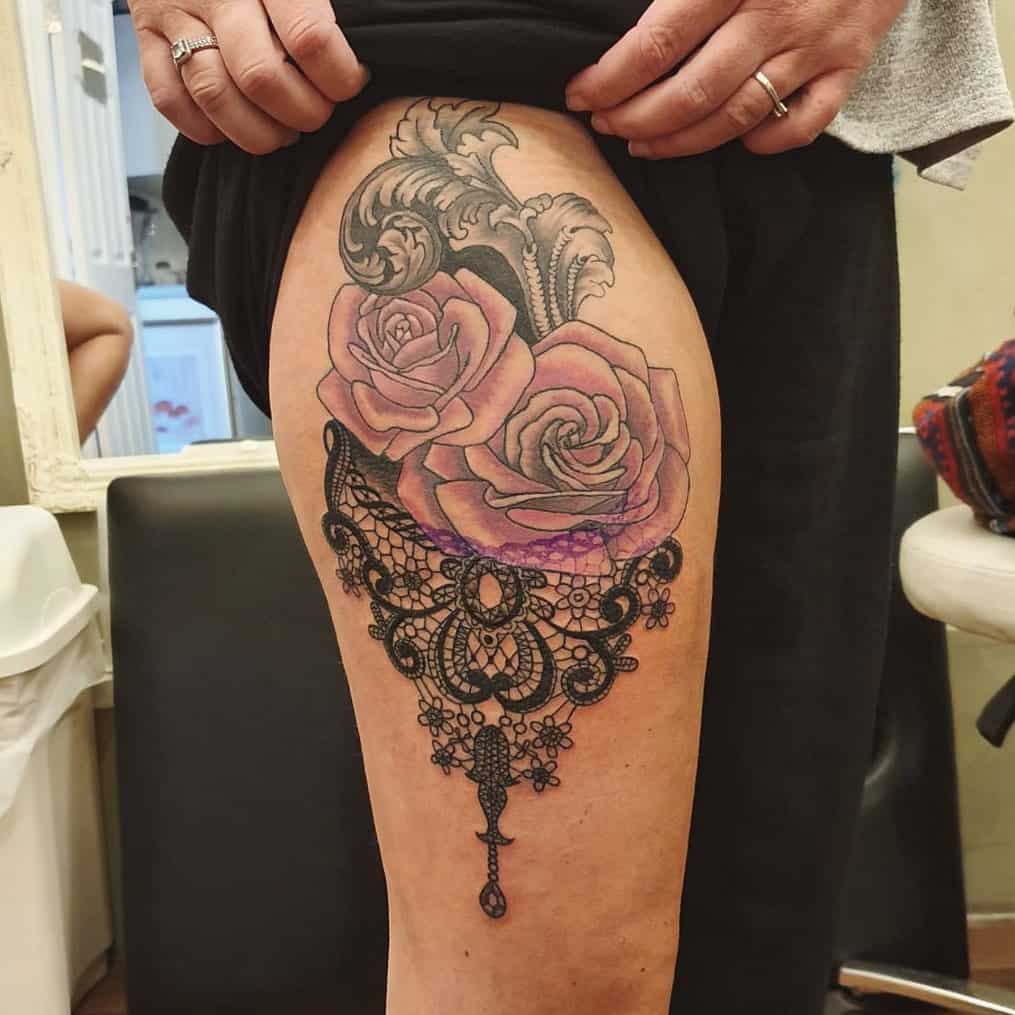 Thigh tattoo featuring detailed pink roses, ornamental filigree, and an intricate black lace design with jewel-like accents.