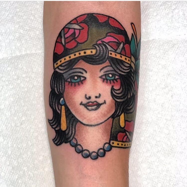 Traditional tattoo of a gypsy woman with bright blue eyes, a floral headscarf, gold earrings, and a pearl necklace on the forearm.