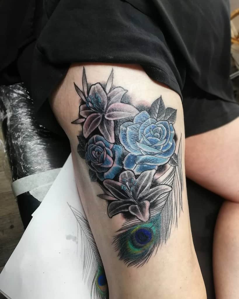 Thigh tattoo featuring blue roses and lilies with black and grey shading, complemented by a detailed peacock feather with vibrant eye colors.