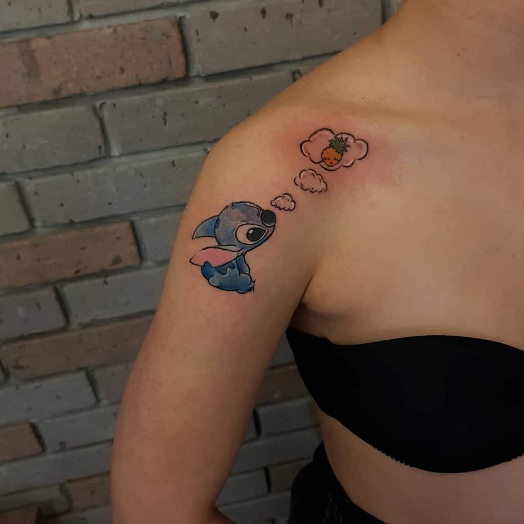 Colorful tattoo of Stitch from Lilo & Stitch thinking of a pineapple, with a cloud-shaped thought bubble, on the shoulder.