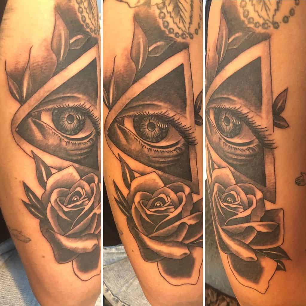 Tattoo of an all-seeing eye inside a triangle, surrounded by roses and leaves, inked in black and grey shading on the forearm.