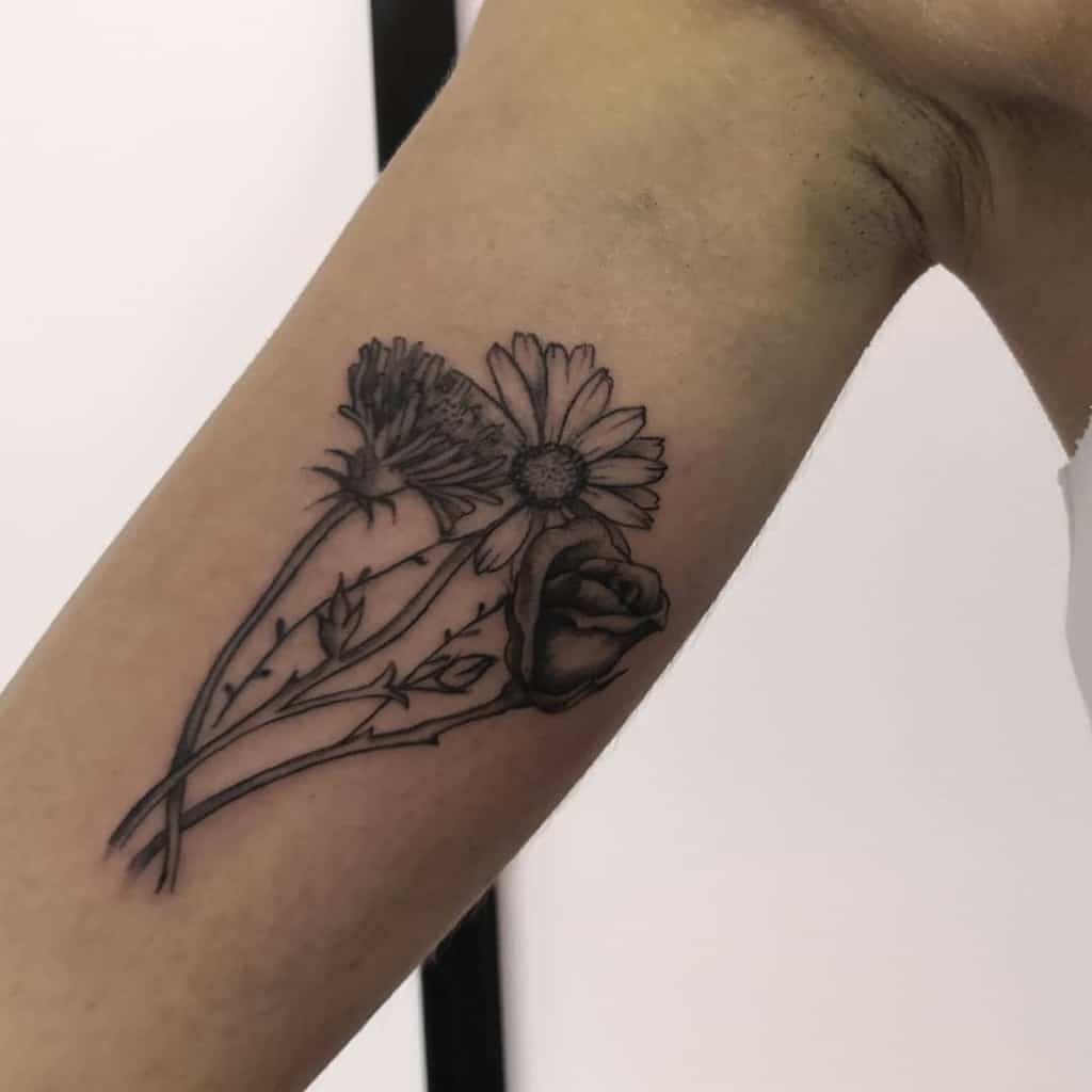 This tattoo features a delicate arrangement of flowers, including a dandelion, a daisy, and a rose, done in fine lines and black ink.