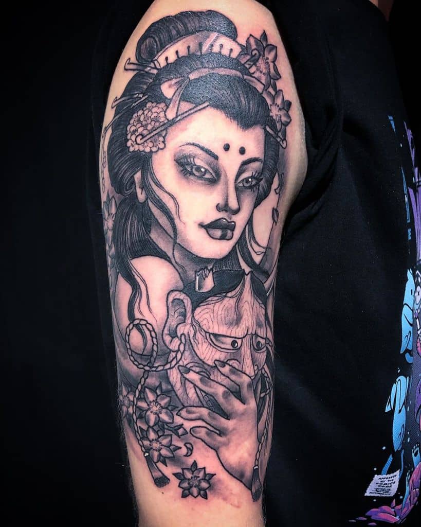 Black and gray upper arm tattoo of a Geisha with intense eyes, intricate floral hair ornaments, and a detailed mask in her hand, blending realism and fantasy.