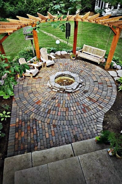 Brick patio features a fire pit, wooden chairs, swing, and pergola in a charming garden setting