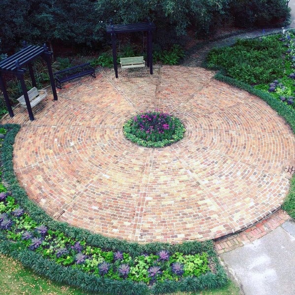 Circular brick patio with benches, pergolas, and central flower bed, embraced by lush greenery
