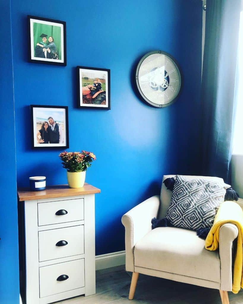 Cozy living room corner with a royal blue wall, beige armchair, framed photos, and bright accents.