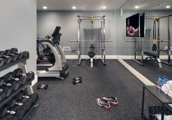 Home gym with rubber flooring, dumbbells, exercise bike, weight rack, and a wall-mounted TV.