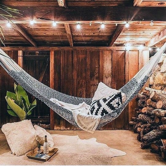 Cozy hammock in a rustic wooden room with string lights, cushions, and stacked firewood.