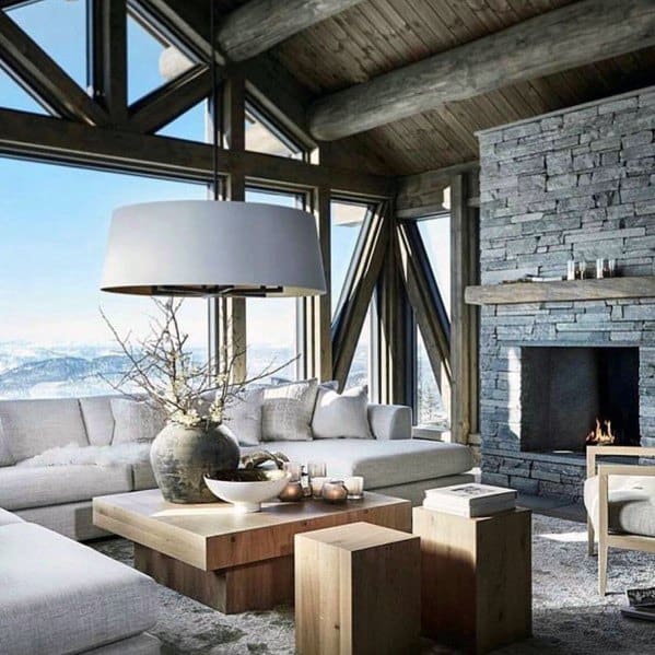 Rustic living room with exposed log beams, stone fireplace, large windows, and cozy white furniture.