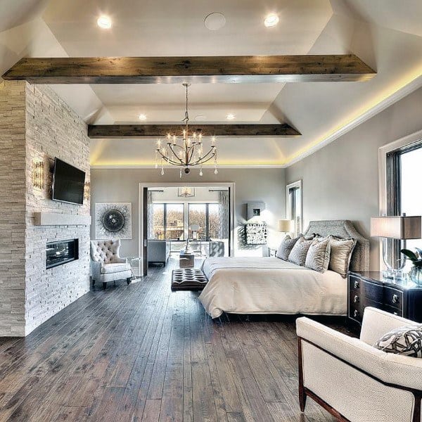 Spacious modern bedroom with wood floors, exposed beams, stone fireplace, and elegant chandelier.