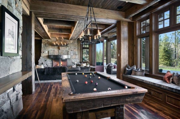 Rustic billiard room with wooden beams, stone fireplace, and large windows overlooking nature.