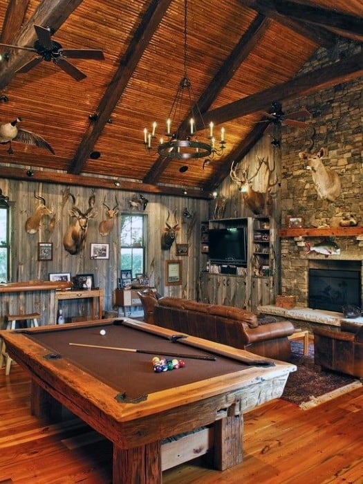 Rustic man cave with wooden beams, pool table, leather couch, stone fireplace, and mounted animal trophies