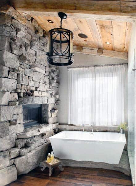 Rustic bathroom with stone walls, wooden ceiling, and large bathtub under a cozy chandelier