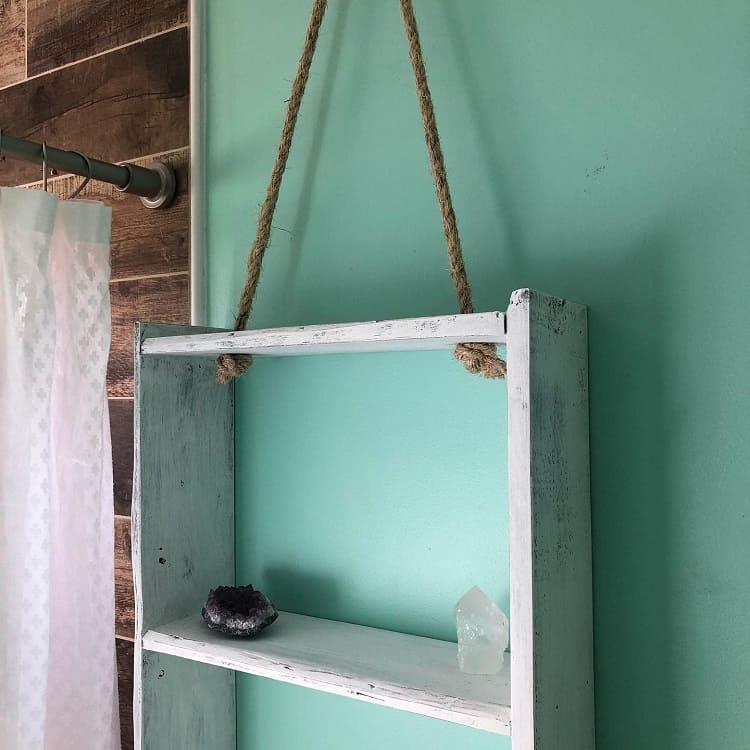 Hanging rustic white wooden bathroom shelf with crystals against teal wall.