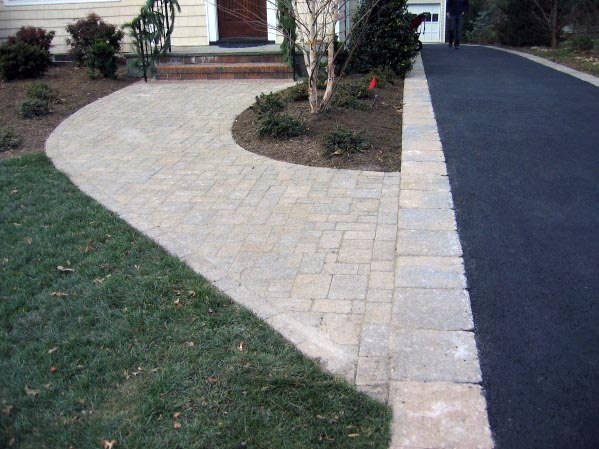 Asphalt driveway with brick edging next to a stone walkway leading to a house entrance.