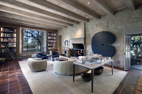 Rustic home library with built-in bookcases, exposed wooden beams, stone walls, and a cozy fireplace, complemented by modern furniture and large windows that bring in natural light.