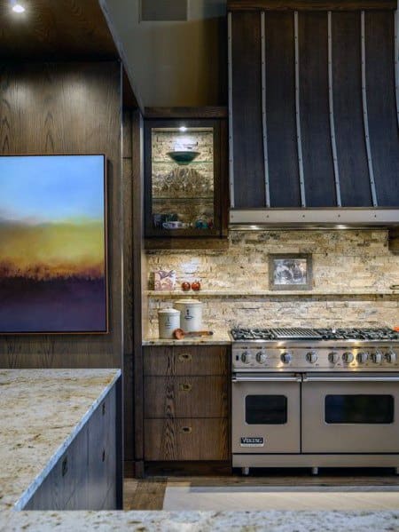 Rustic kitchen with a warm stone backsplash, dark wood cabinetry, stainless steel range, and ambient accent lighting