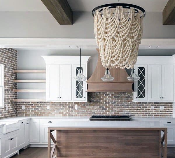 Rustic kitchen with brick backsplash, wooden island, white cabinets, and a beaded chandelier.