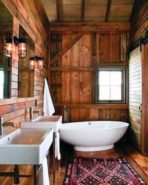 Rustic bathroom with wooden walls, white bathtub, dual sinks, and a patterned rug