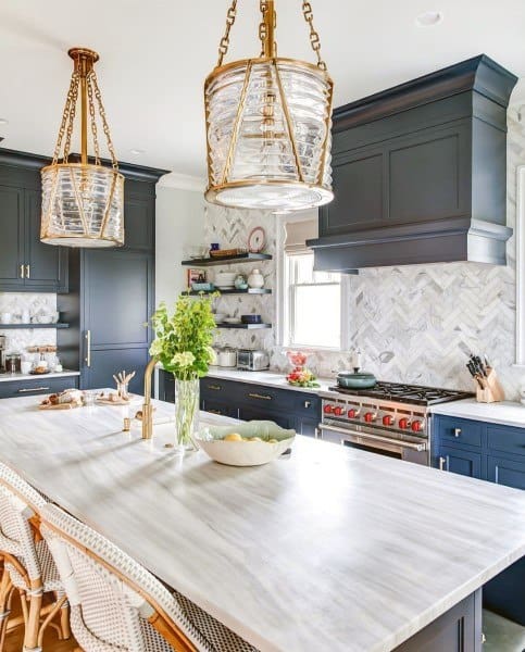 Navy kitchen cabinets with marble chevron backsplash, brass lighting, and large island countertop.