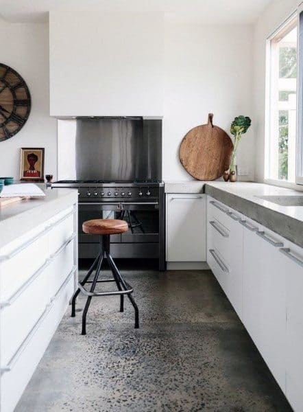 Modern kitchen with white cabinets, metal stool, stainless steel oven, and concrete floor. Large window brightens the space