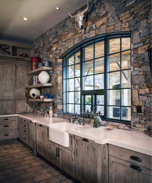 Rustic kitchen with stone walls, arched window, wooden cabinets, and a farmhouse sink.