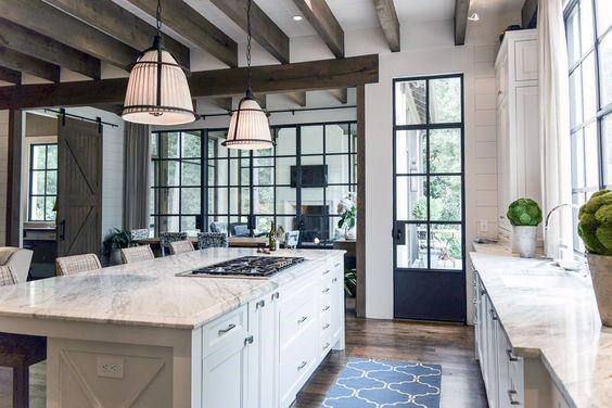 Bright kitchen with white cabinets, marble countertops, wooden beams, and large windows.