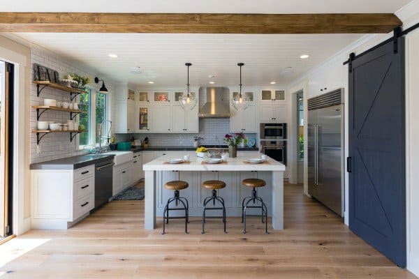 Bright rustic kitchen featuring a white island, wood stools, barn door, and exposed ceiling beam.