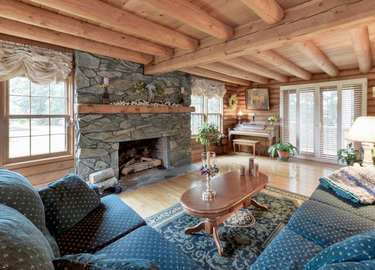 Cozy log cabin living room with stone fireplace, wooden beams on the ceiling, sofas, and a large window